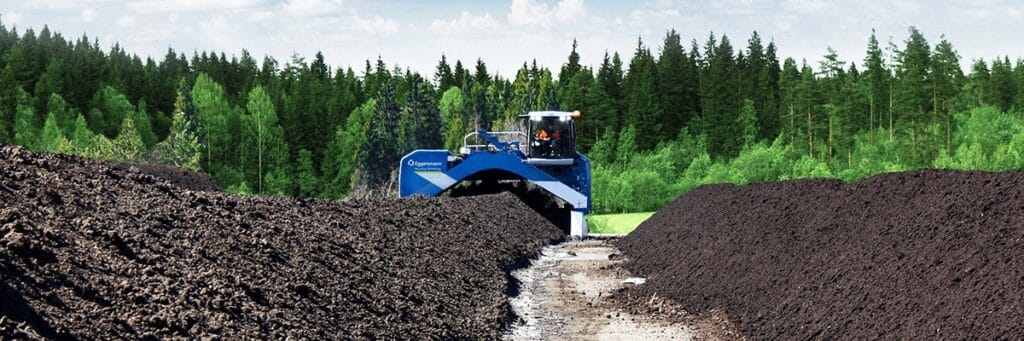 Retourneur d'andains Eggermann traitant des tas de compost sombre devant une forêt dense de conifères.