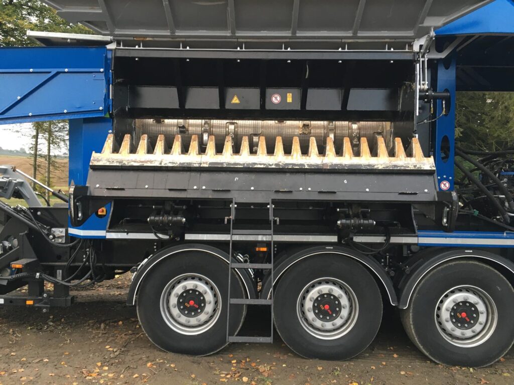 Broyeur forestier ou déchiqueteuse mobile sur camion lourd bleu et noir, vues des roues et du tambour d'alimentation denté. Panneaux d'avertissement visibles.
